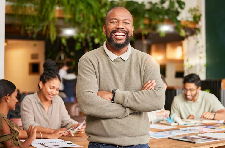 A confident black man smiles at the camera while his employees work on a project behind him
